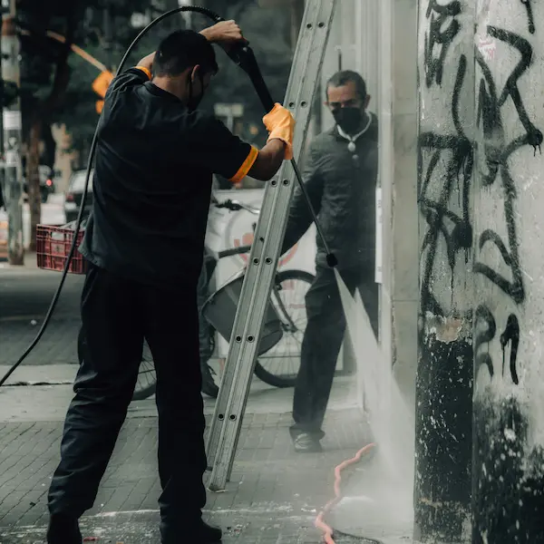 Graffiti Removal Pressure Washing showing a worker pressure washing a storefront wall with graffiti and surface buildup.